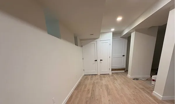 Empty finished basement with light wood flooring, white walls, two closed white doors with black handles, and recessed ceiling lights.