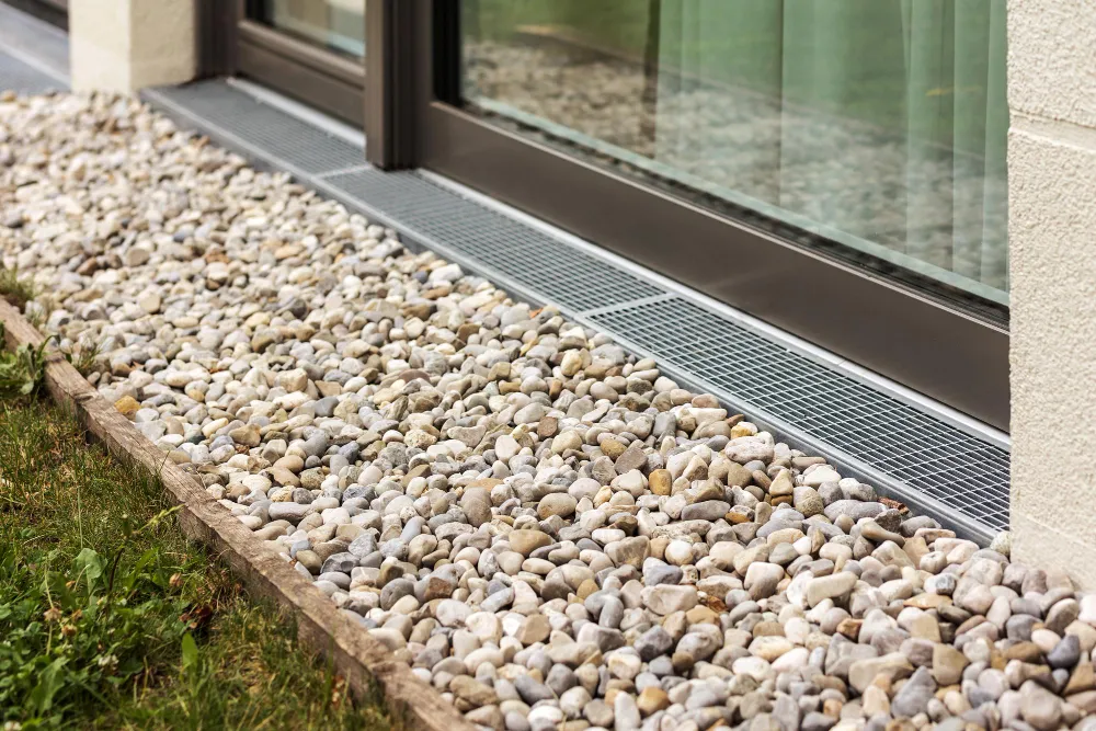 Metal drainage grate installed along the edge of a building foundation with a bed of small, light-colored stones surrounding it, bordered by a strip of grass.