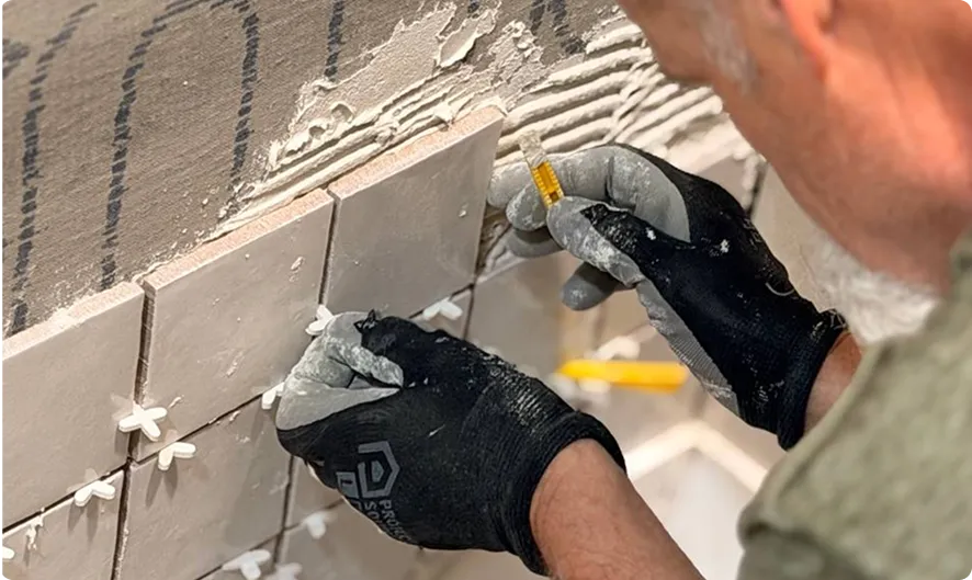 Close-up of a man wearing black gloves installing square wall tiles with spacers on a wall, applying adhesive with a small tool.