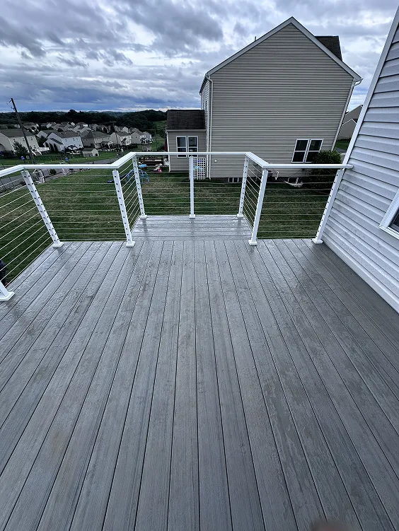 View of a clean, gray wooden deck with sleek white metal railings and cable wires, overlooking a suburban neighborhood with houses, yards, and a cloudy sky.