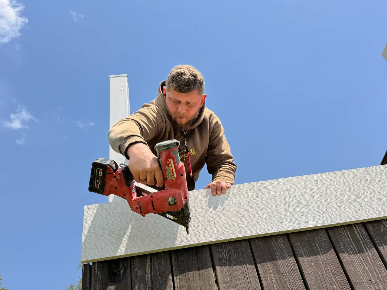 Man in a brown hoodie using a red nail gun to attach a white board to a wooden fence under a clear blue sky.