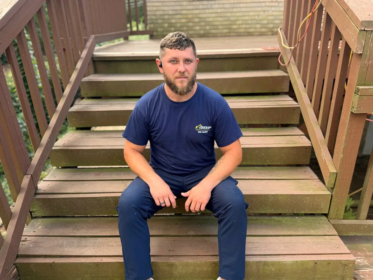 Man with beard wearing a navy blue X-GROUP t-shirt and dark pants sitting on wooden steps outdoors, looking directly at the camera.