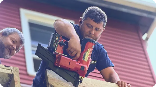 Man using a red nail gun to secure wooden planks on an outdoor structure, with another man watching nearby and a red building with white trim in the background.