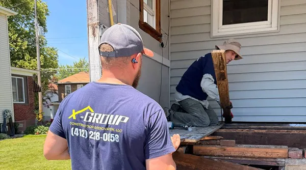 Two construction workers renovating a wooden deck of a house; one is kneeling inside the porch area holding a wooden plank, and the other is standing outside wearing a cap and a blue t-shirt with 'X-GROUP Construction & Renovation LLC' and a phone number printed on the back.