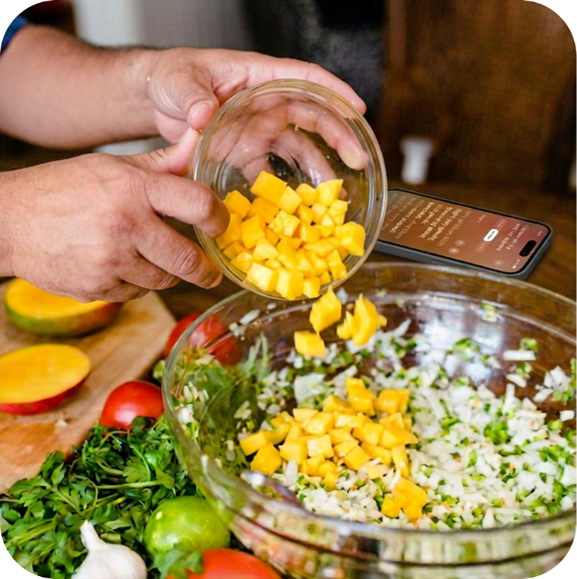 Hands adding diced mango to a bowl of salsa beside fresh ingredients and a phone