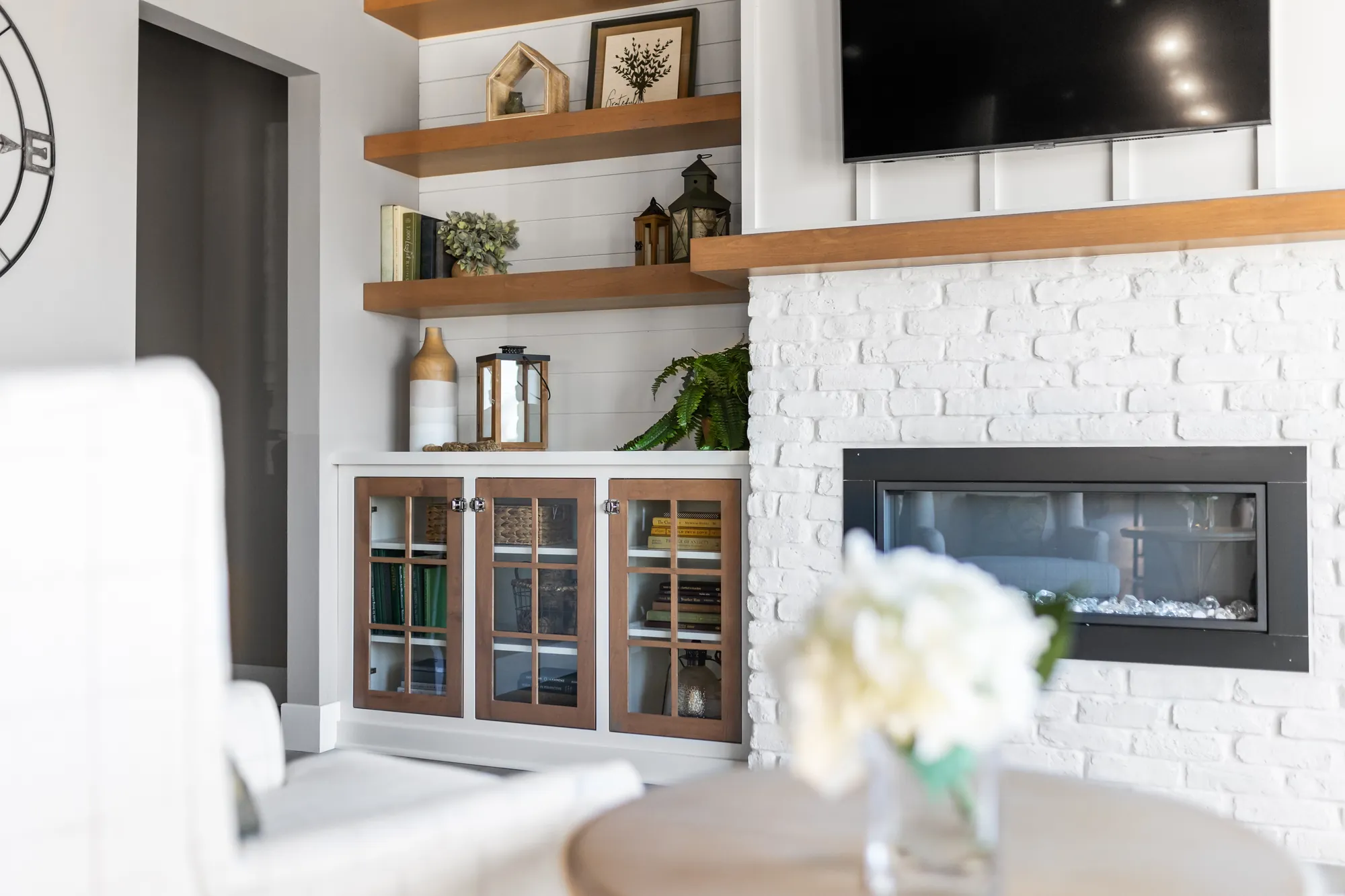 White brick fireplace with a wood mantel, floating shelves and glass-front cabinets