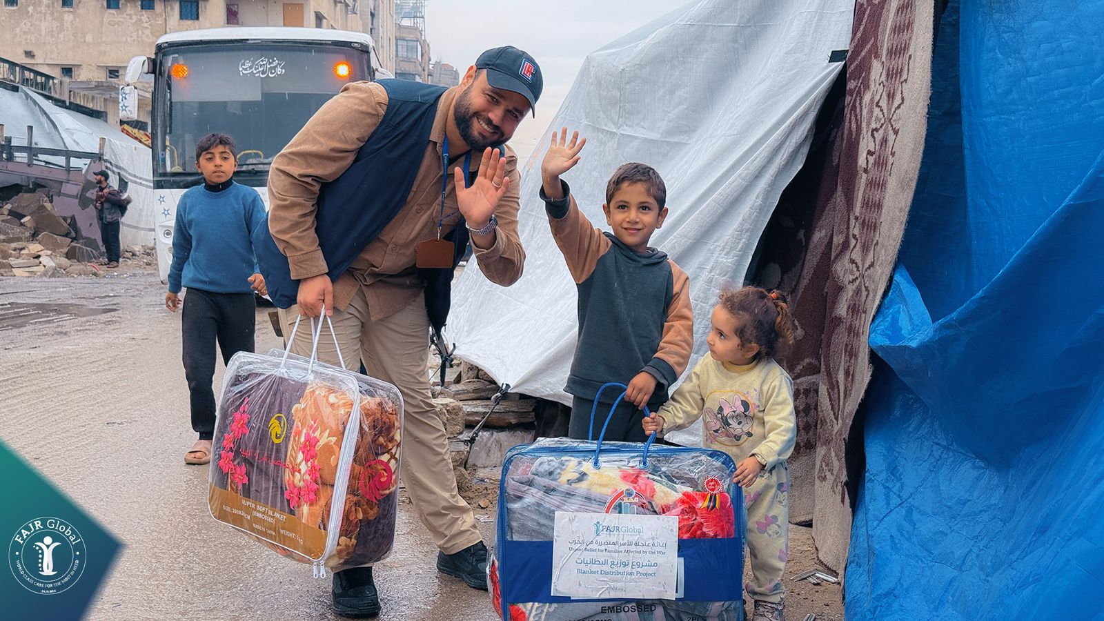 A FAJR Global volunteer waves with two children beside winter blanket bags in Gaza