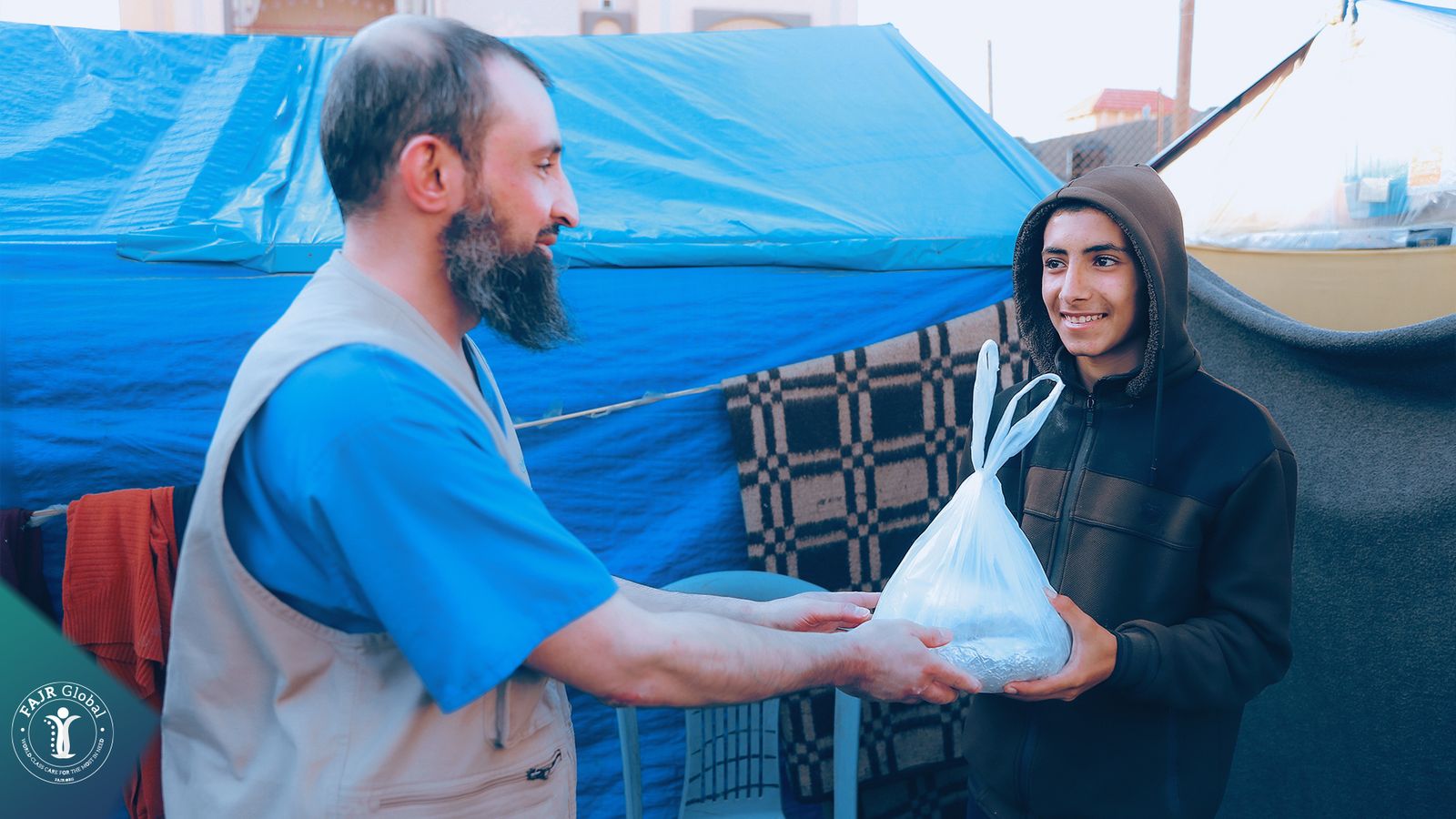 A FAJR aid worker handing a bag of supplies to a young man outside a tent