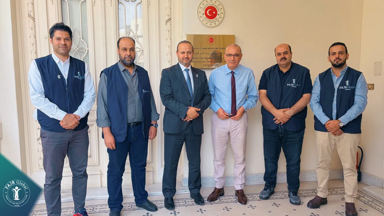 FAJR team members and officials standing beside the Turkish Embassy emblem on a wall