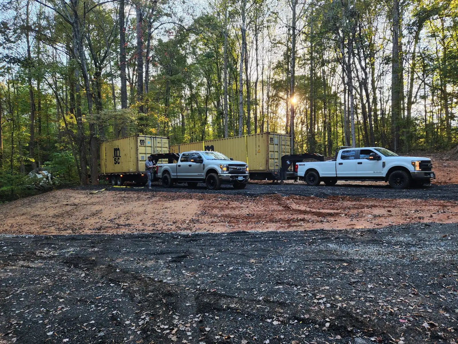 Two white pickup trucks parked on a gravel and dirt pad in a wooded area with two large yellow shipping containers on trailers behind them, with a person standing near one truck.