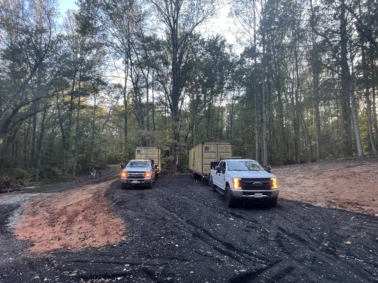 Two Central Virginia Containers pickup trucks on a dirt lane through wooded property during a container delivery