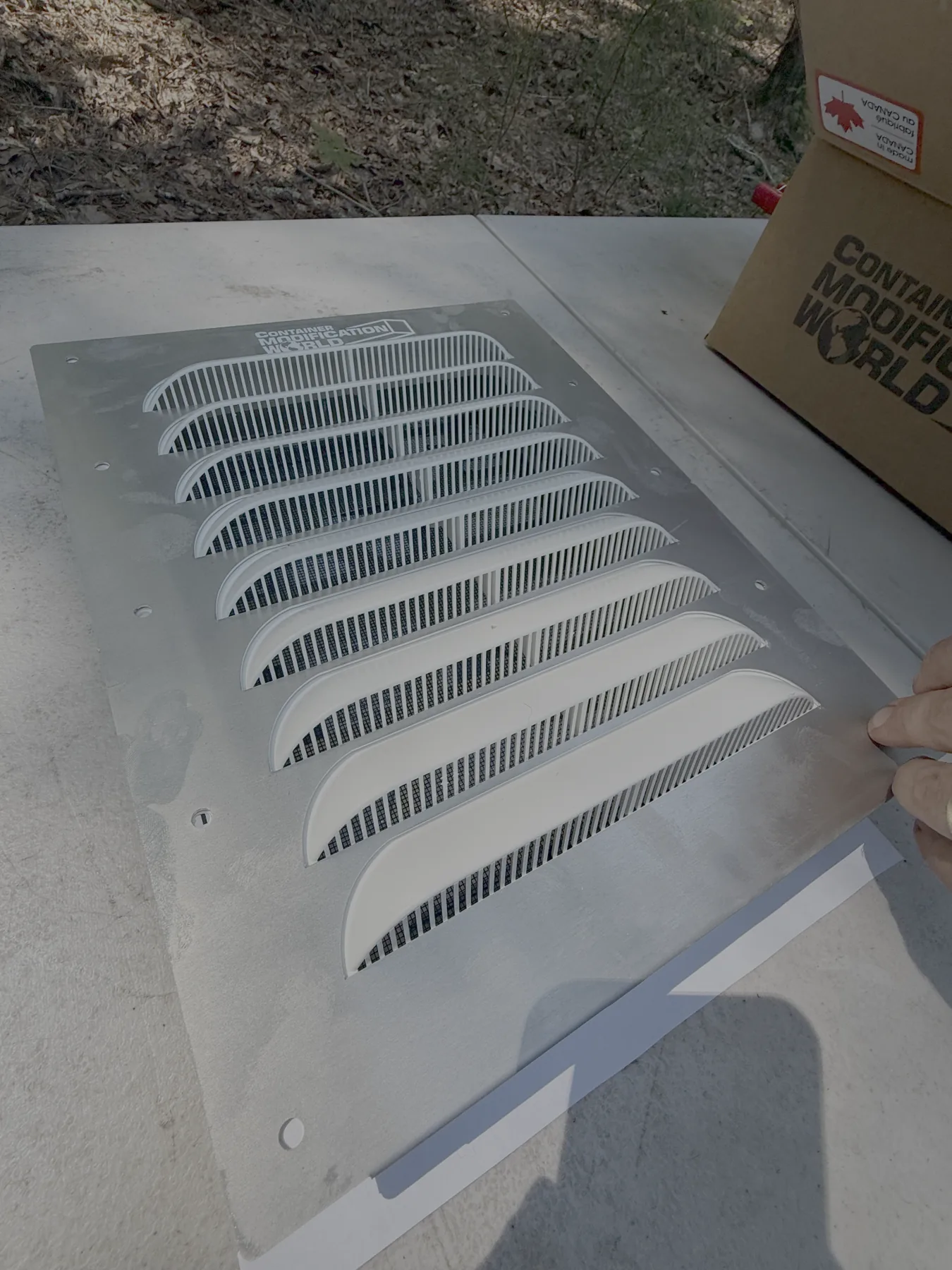 Rectangular metal vent panel with angled slats, held by a hand on a concrete surface with a cardboard box labeled "Container Modification World" nearby.