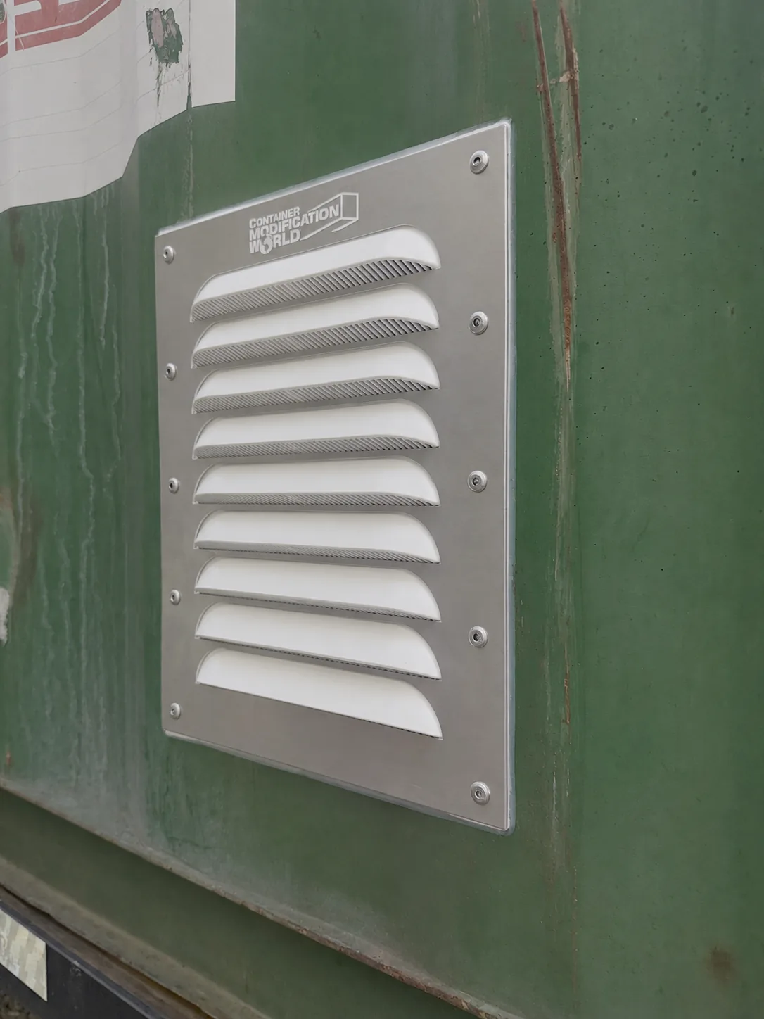 Closeup of a metallic ventilation panel with horizontal slats mounted on a weathered green surface, featuring the text 'CONTAINER MODIFICATION WORLD' at the top left.