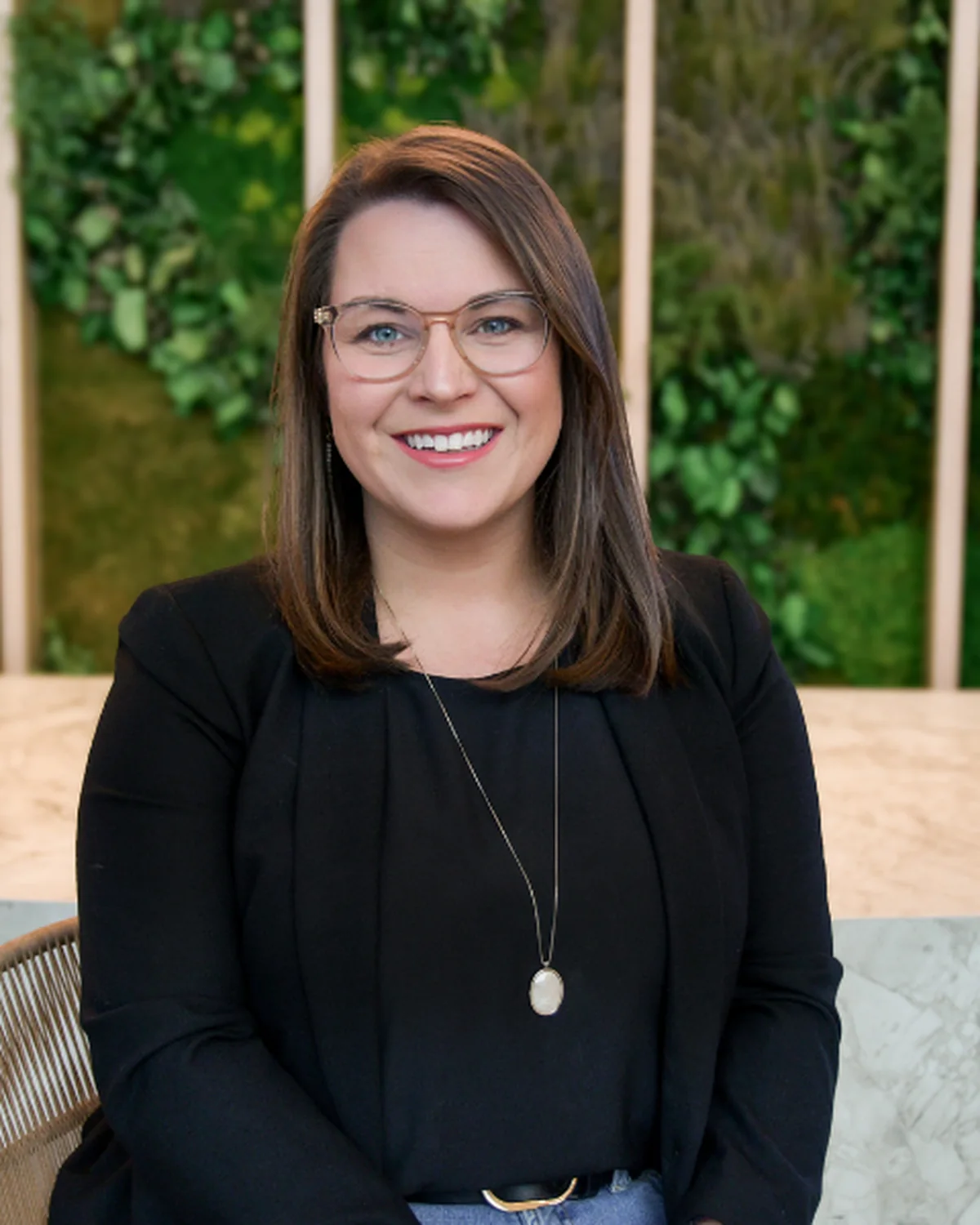 Smiling woman with shoulder-length brown hair wearing glasses, a black blazer, black top, long necklace, and blue jeans, sitting indoors with a green leafy background.