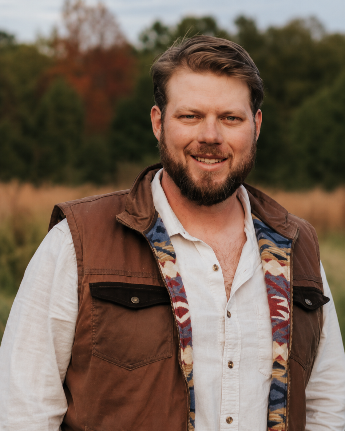 Smiling man with short brown hair and beard wearing a brown jacket with a patterned lining over a white buttoned shirt, standing outdoors with blurred trees and grass in the background.