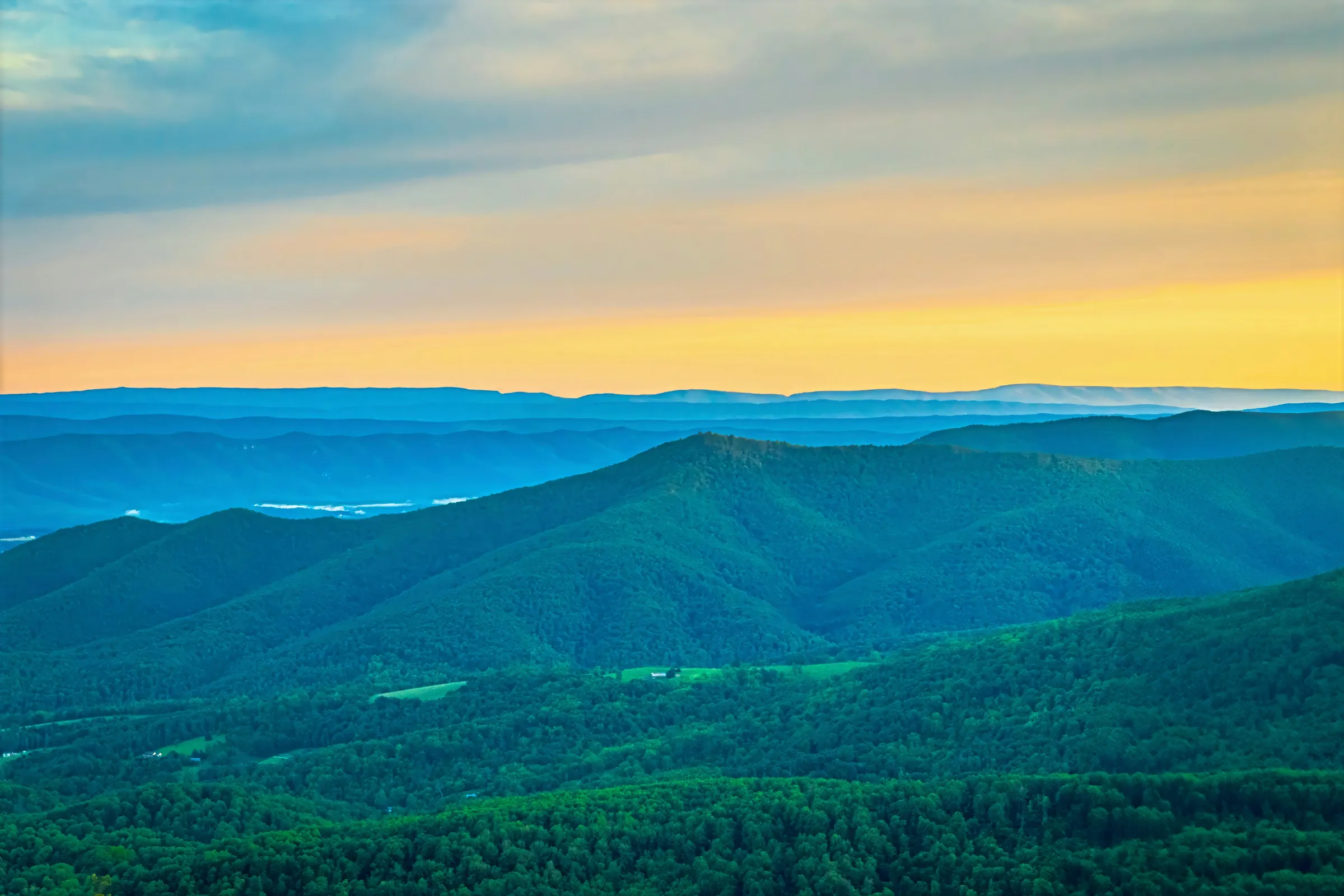 View from the Blue Ridge Parkway in Virginia: layers of forested mountain ridges under a golden evening sky, with small farm fields in the valley below