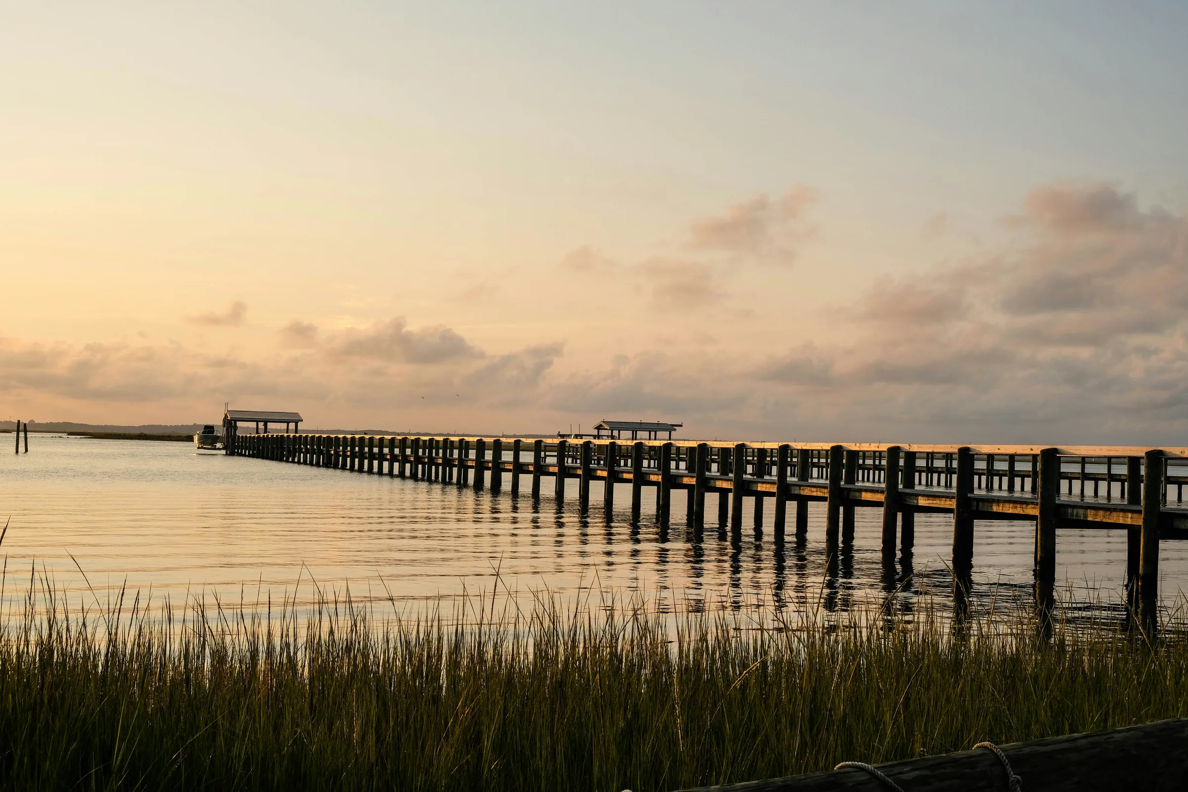 A long wooden pier with covered boat lifts stretching across calm water at sunset, with marsh grass in the foreground