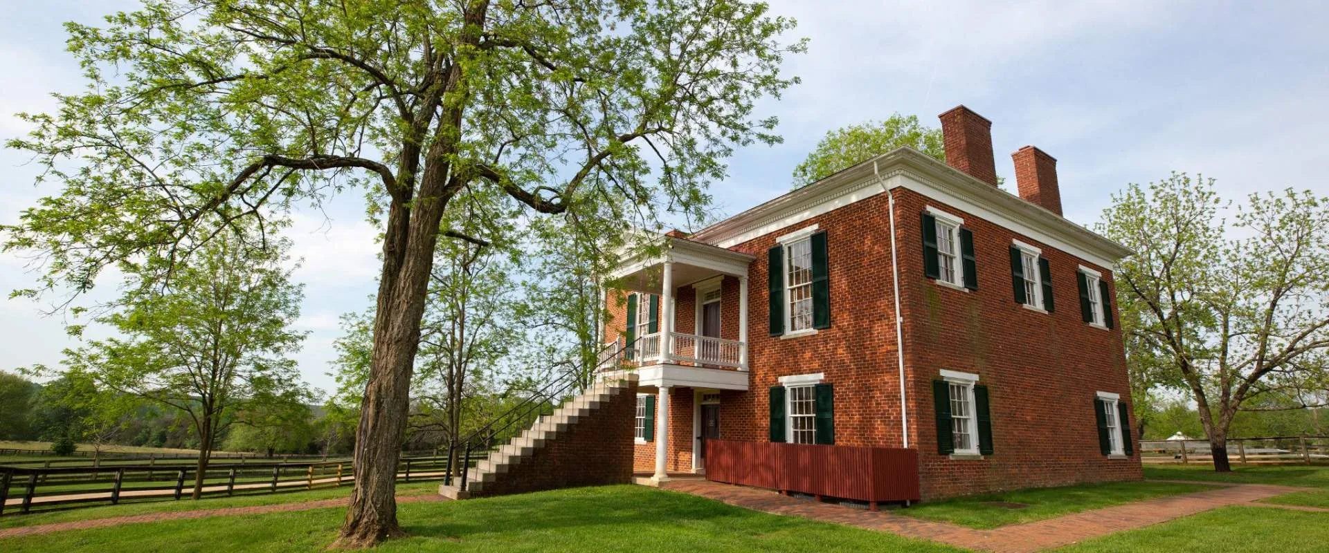 Red brick courthouse with green shutters and an outside staircase at Appomattox Court House National Historical Park, framed by spring trees and split-rail fences