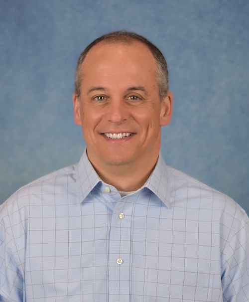 Headshot of a smiling man wearing a blue dress shirt in front of a blue backround