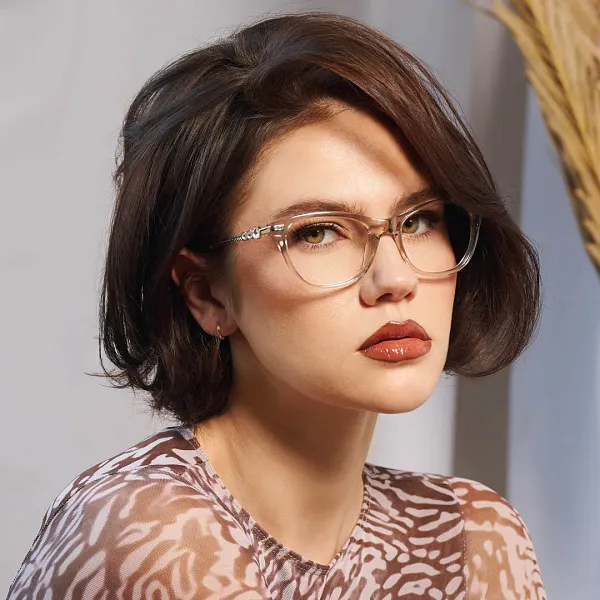 Portrait of a young woman with short dark brown hair wearing clear-framed glasses and glossy brown lipstick, dressed in a patterned top with neutral tones, against a blurred background with beige and blue elements.