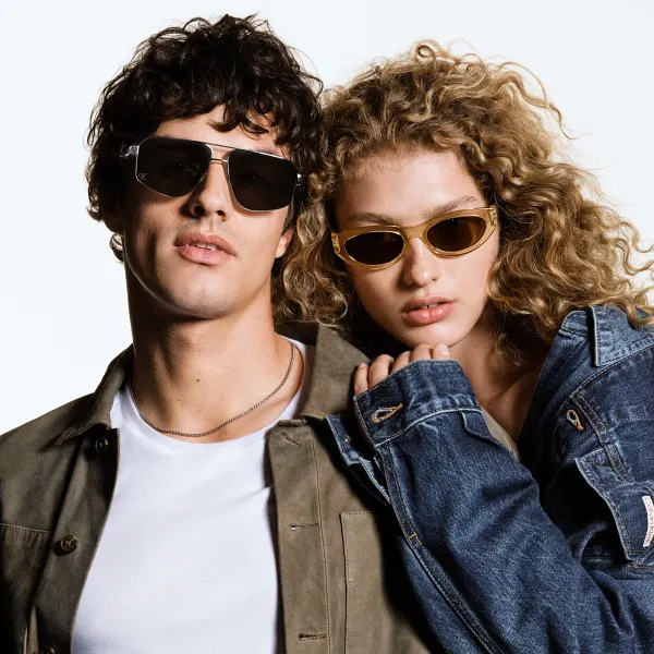 Portrait of a brown haired man wearing a brown jacket and a blonde curly haired woman wearing a denim jacket modeling Calvin Klein Sunglasses.
