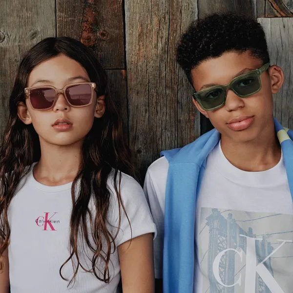 Picture of one boy and one girl standing in front of wood planks wearing Calvin Klein sunglasses.