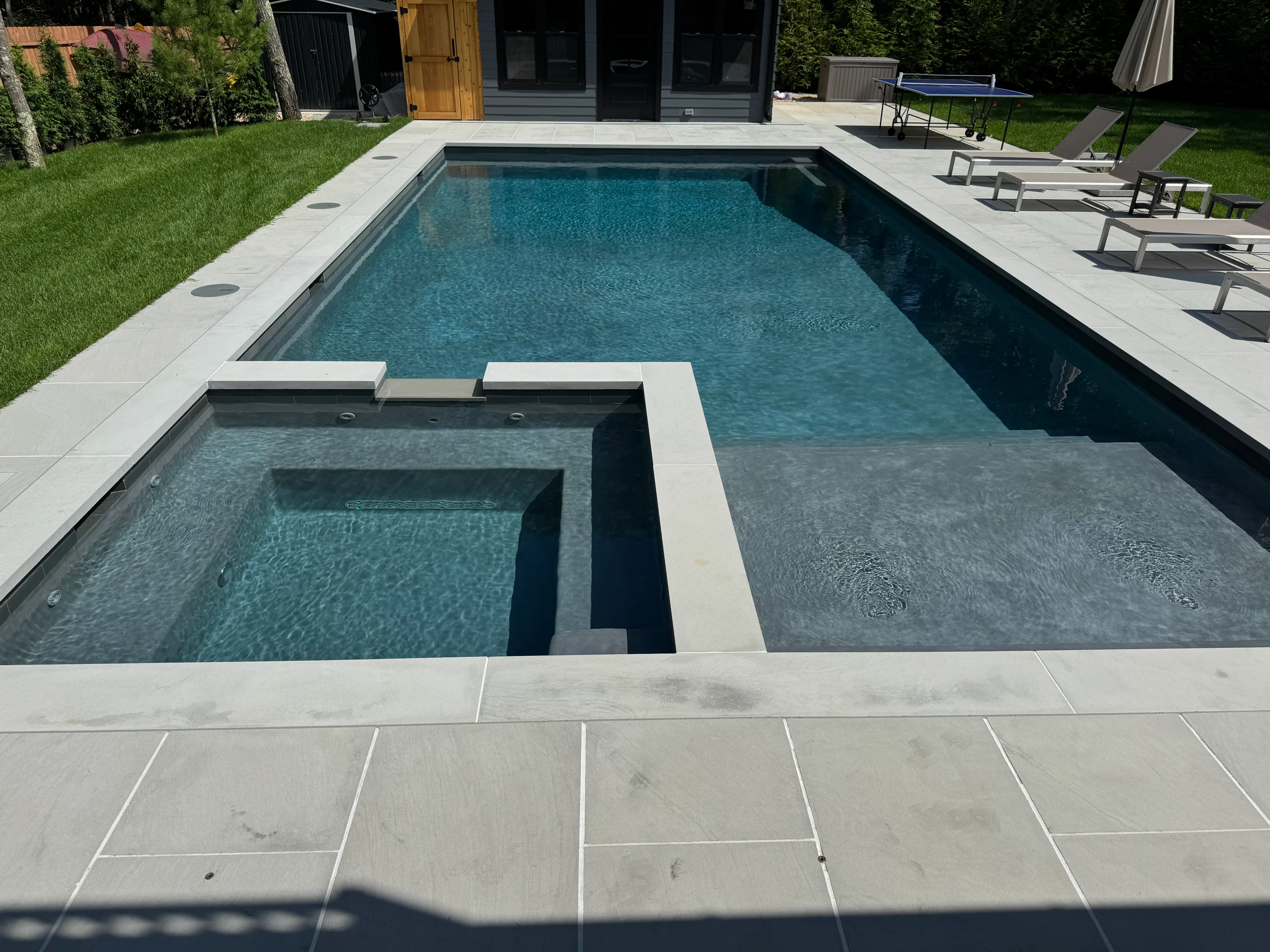Modern in-ground pool with integrated spa, tanning ledge, and light stone patio on Long Island, NY near Hampton Bays.