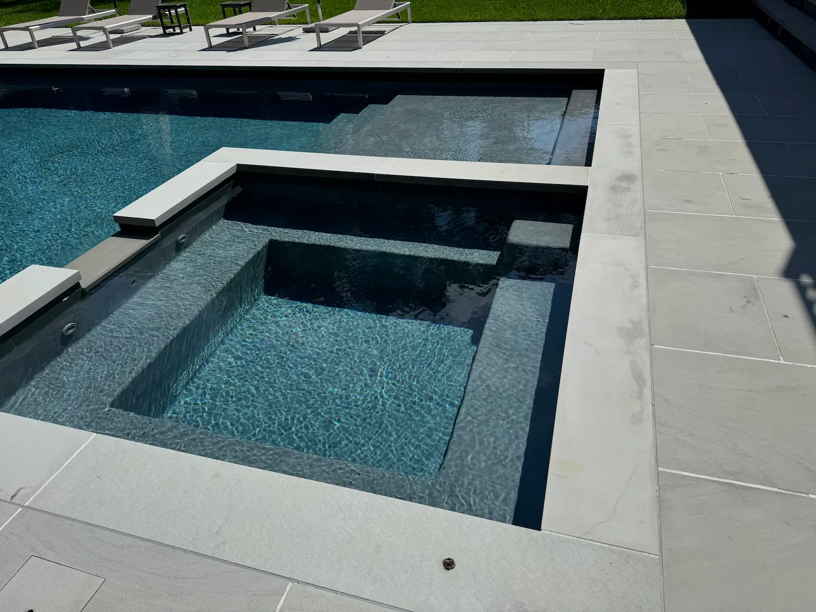 Modern in-ground pool and spa with built-in seating, clear water, and light stone decking on Long Island, NY near Westhampton Beach.