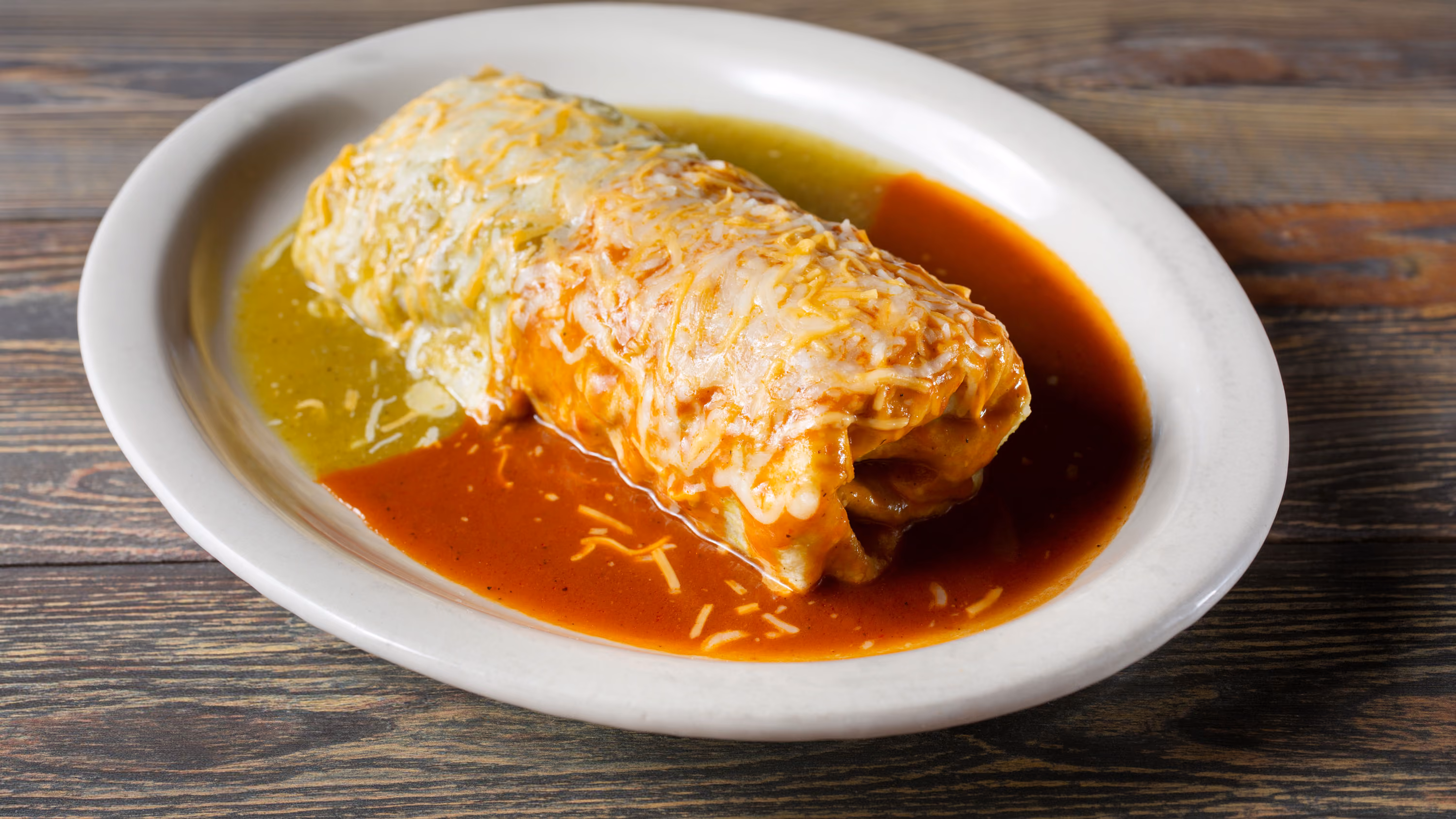 A large burrito covered with melted cheese and red and green sauce on a white oval plate.