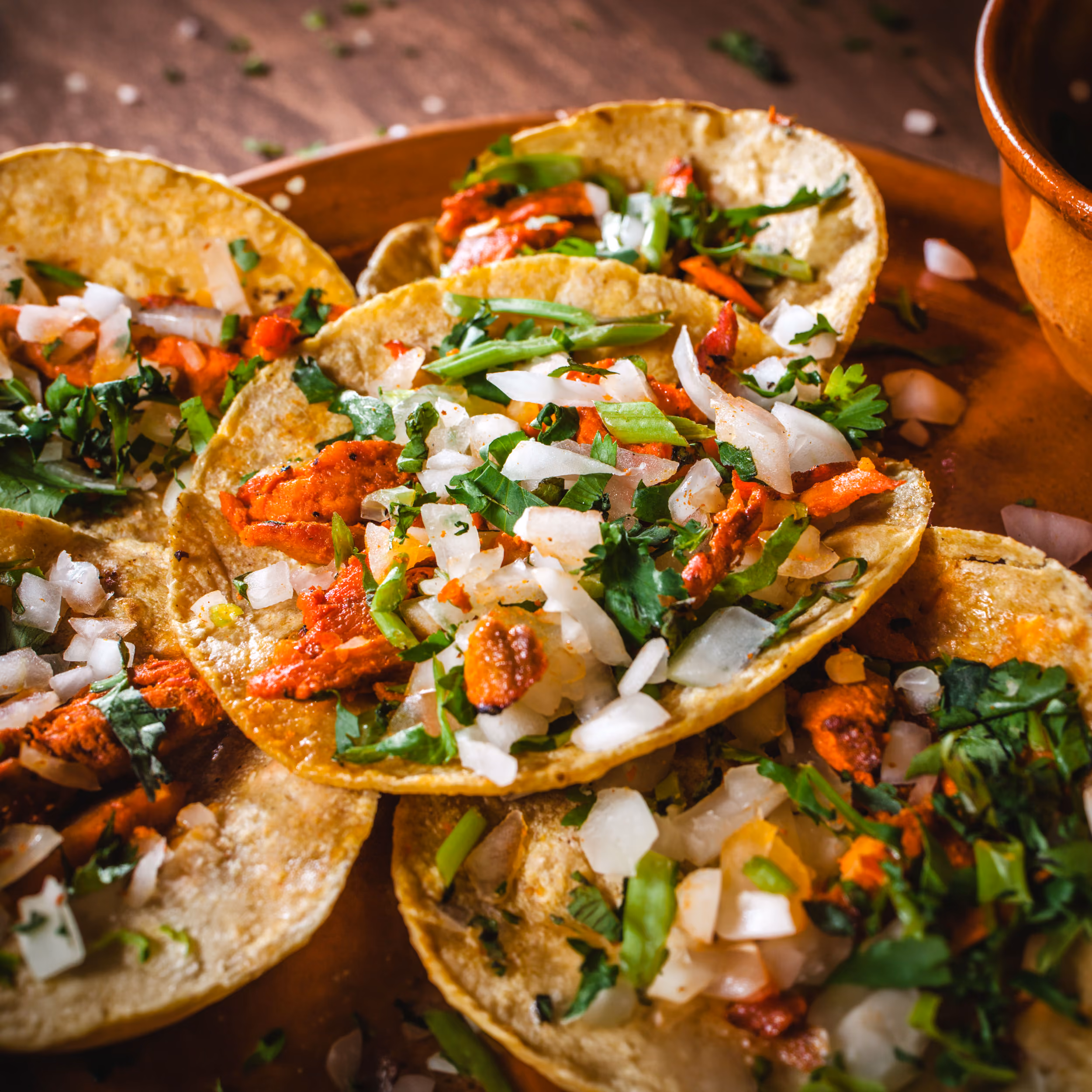 Close-up of tacos with grilled meat, chopped onions, cilantro, and green herbs on a brown plate.