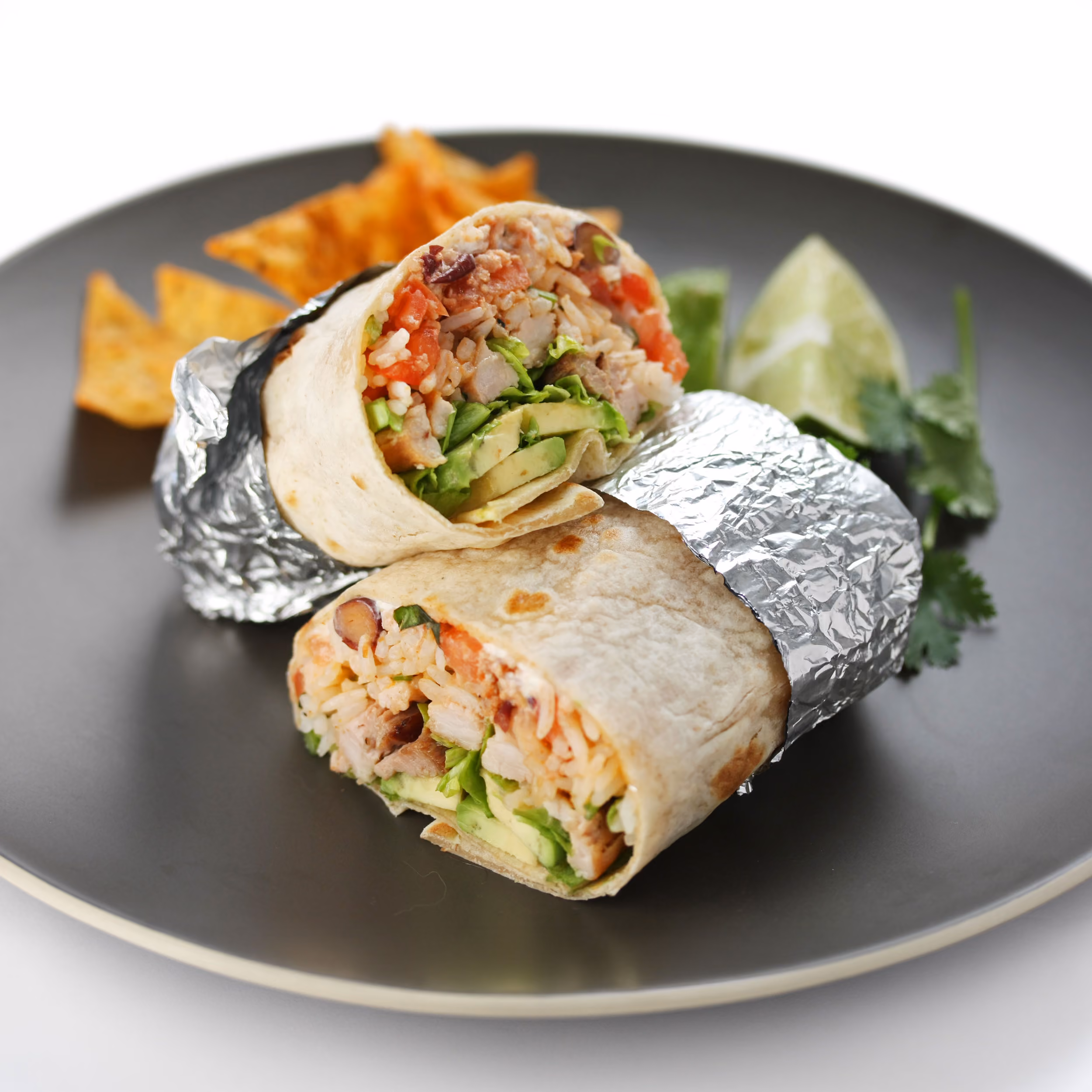 Cut chicken burrito with rice, beans, and avocado wrapped in foil, served with chips and a lime wedge.