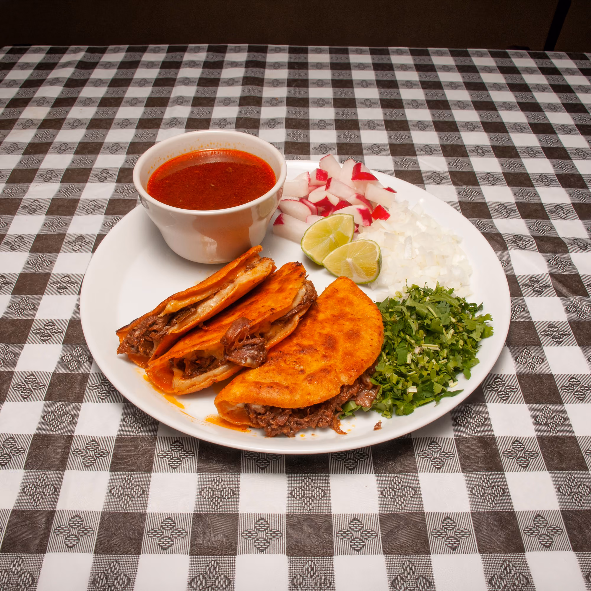 Plate with three quesabirria tacos, chopped onions, cilantro, radishes, lime, and a cup of consome.