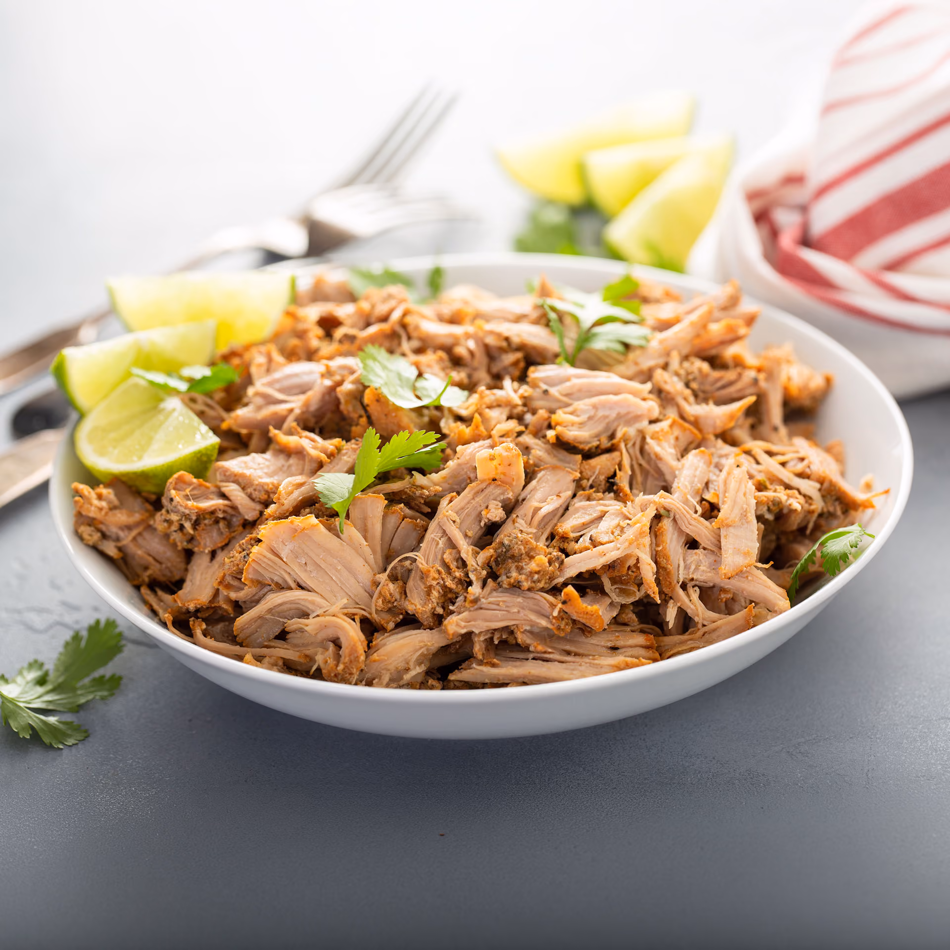 White bowl with shredded carnitas meat garnished with cilantro and lime wedges on a gray surface.