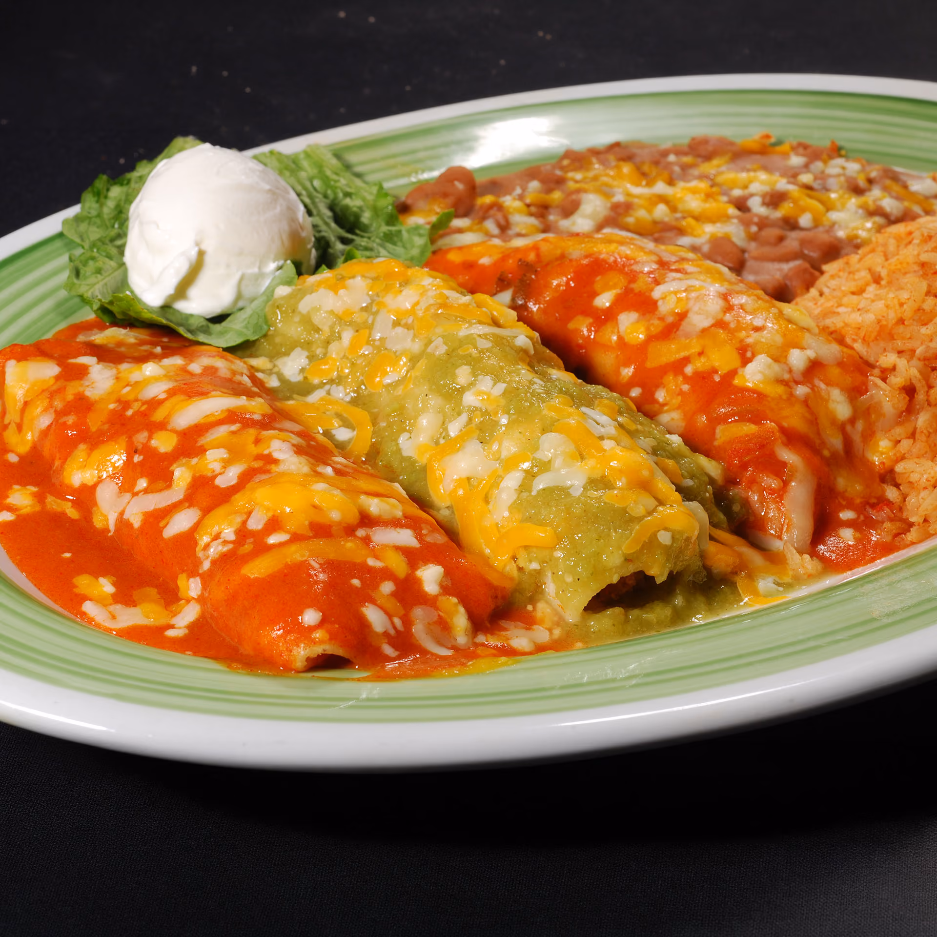 Plate with three cheese-covered enchiladas, refried beans, Mexican rice, lettuce, and sour cream.