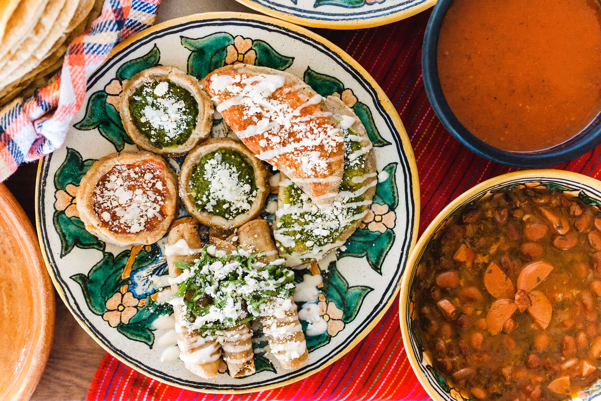 Plate of Mexican snacks with sopes, chalupas, flautas topped with cheese and crema next to beans and red sauce.