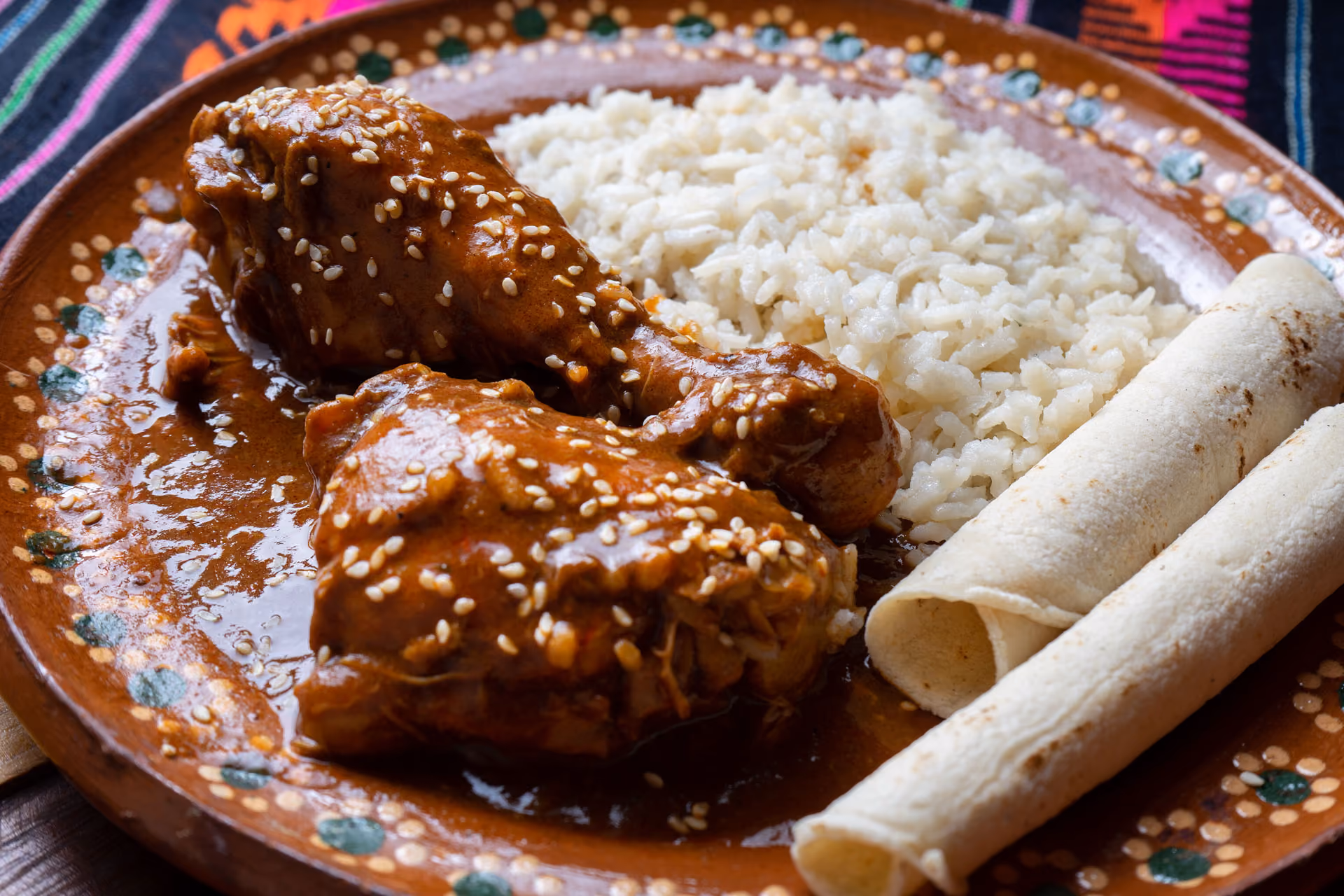 Plate with chicken covered in mole sauce with sesame seeds, white rice, and rolled tortillas.