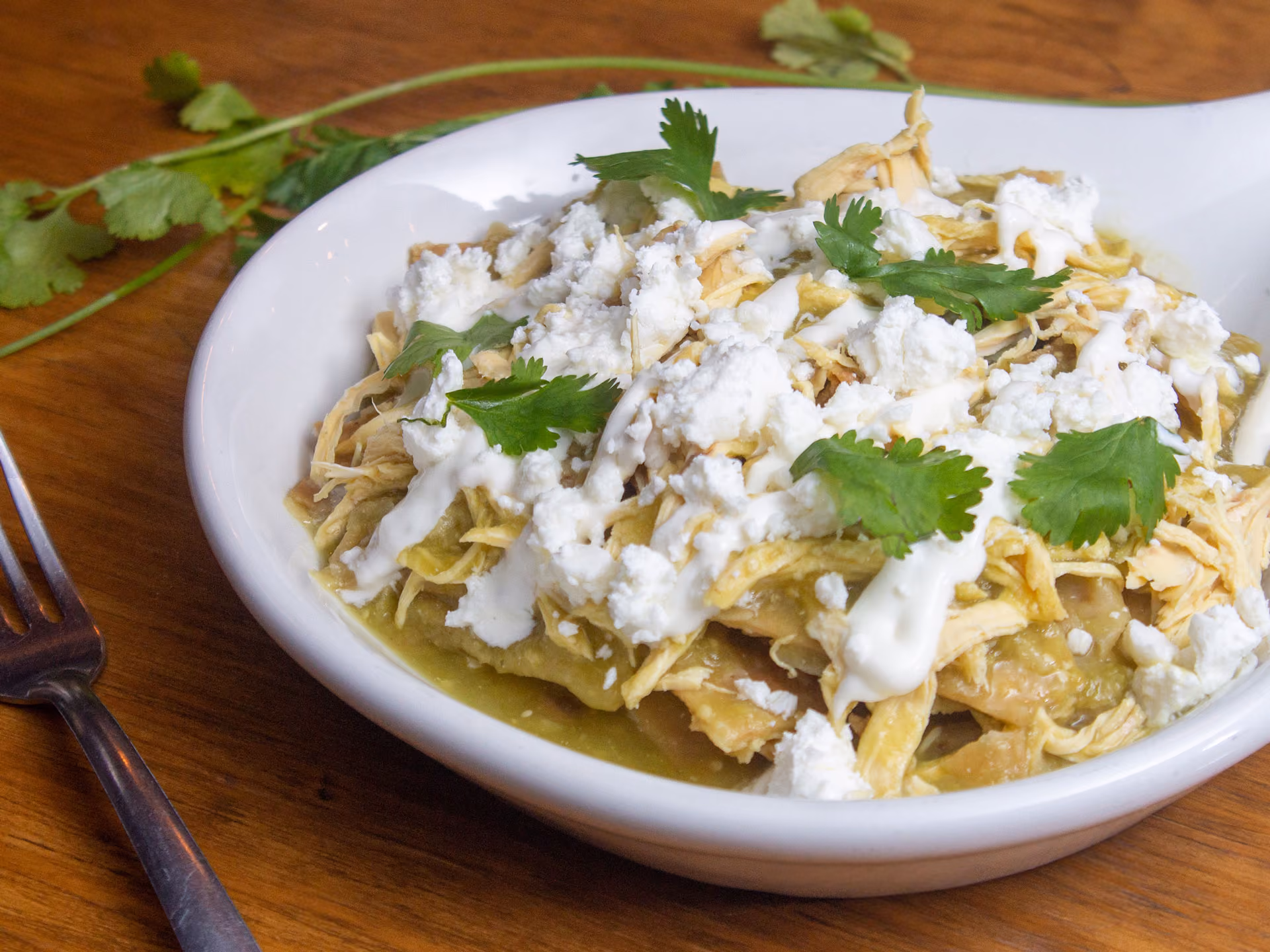 Plate of green chilaquiles topped with shredded chicken, white cheese, sour cream, and cilantro leaves.