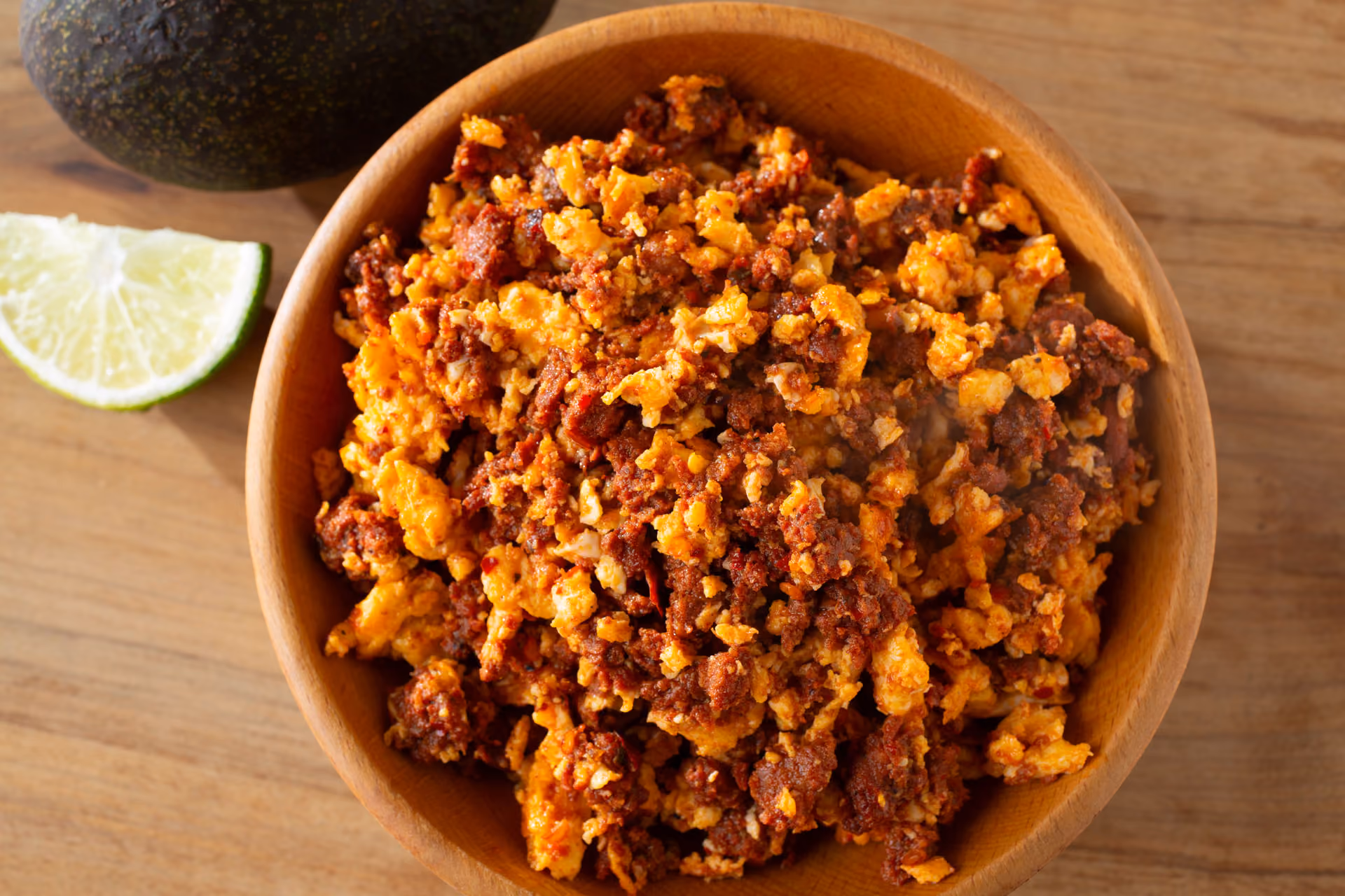 Wooden bowl filled with scrambled eggs and chorizo on a wooden table with avocado and lime slice nearby.