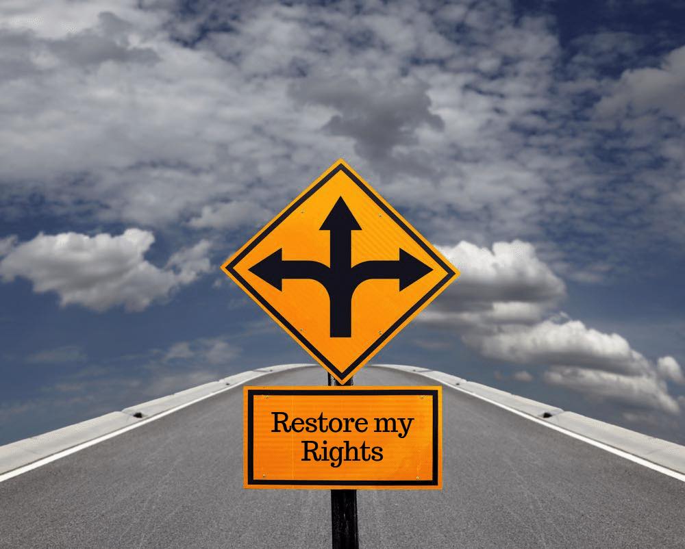Yellow road sign with three arrows and a smaller sign reading 'Restore my Rights' on an empty highway
