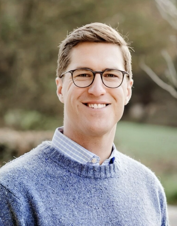Smiling man with glasses wearing a blue sweater and checkered shirt outdoors with blurred background.