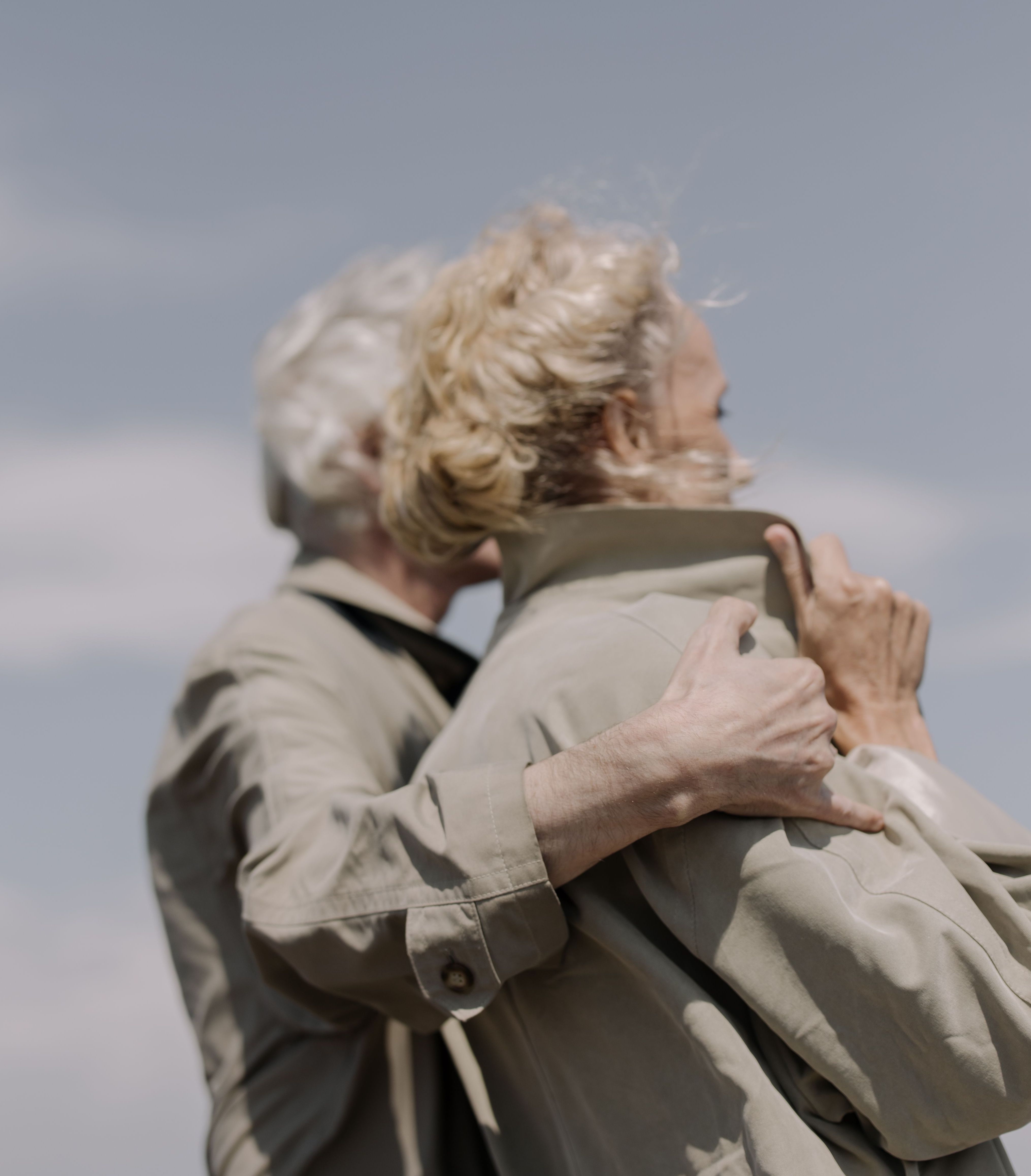Two elderly people in beige coats embracing outdoors with a blue sky background.