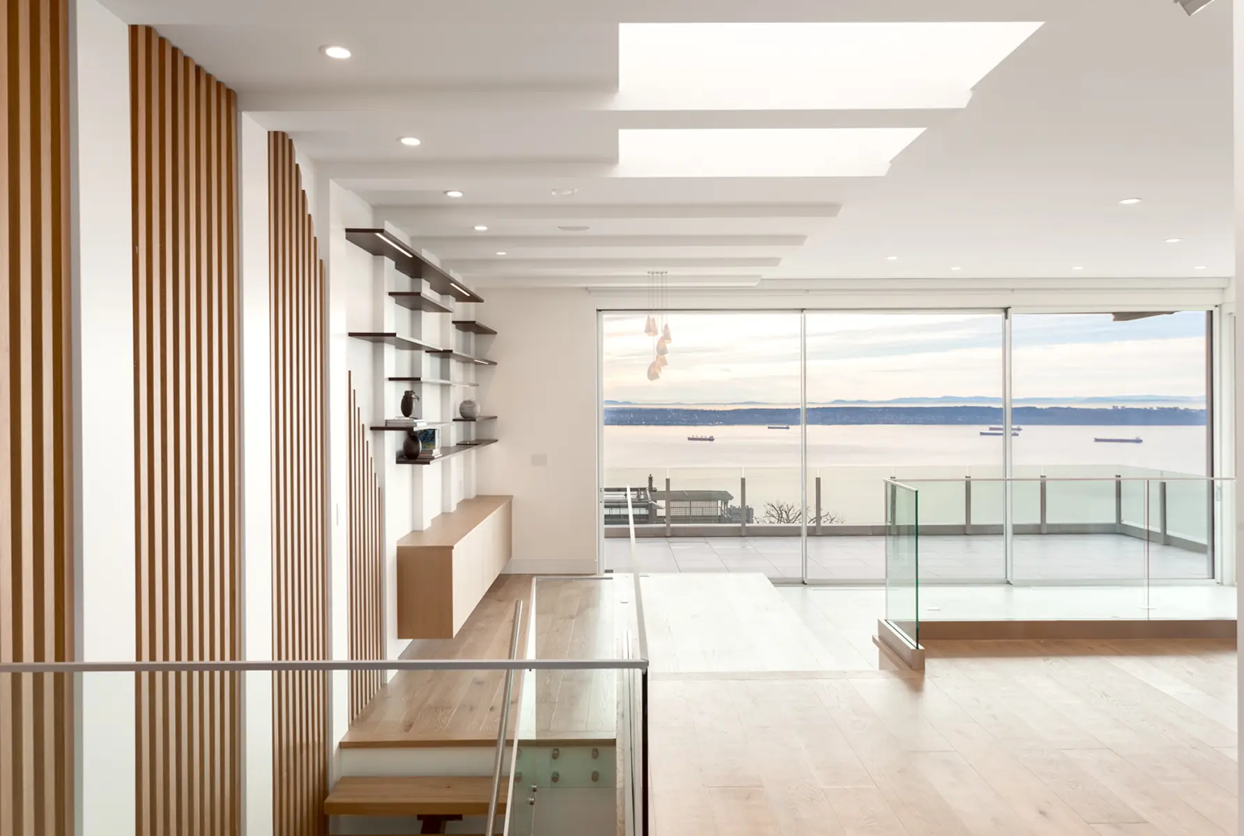 Open living space with a slatted wood divider, floating shelves and a glass balcony facing the water, Folkstone Way rebuild
