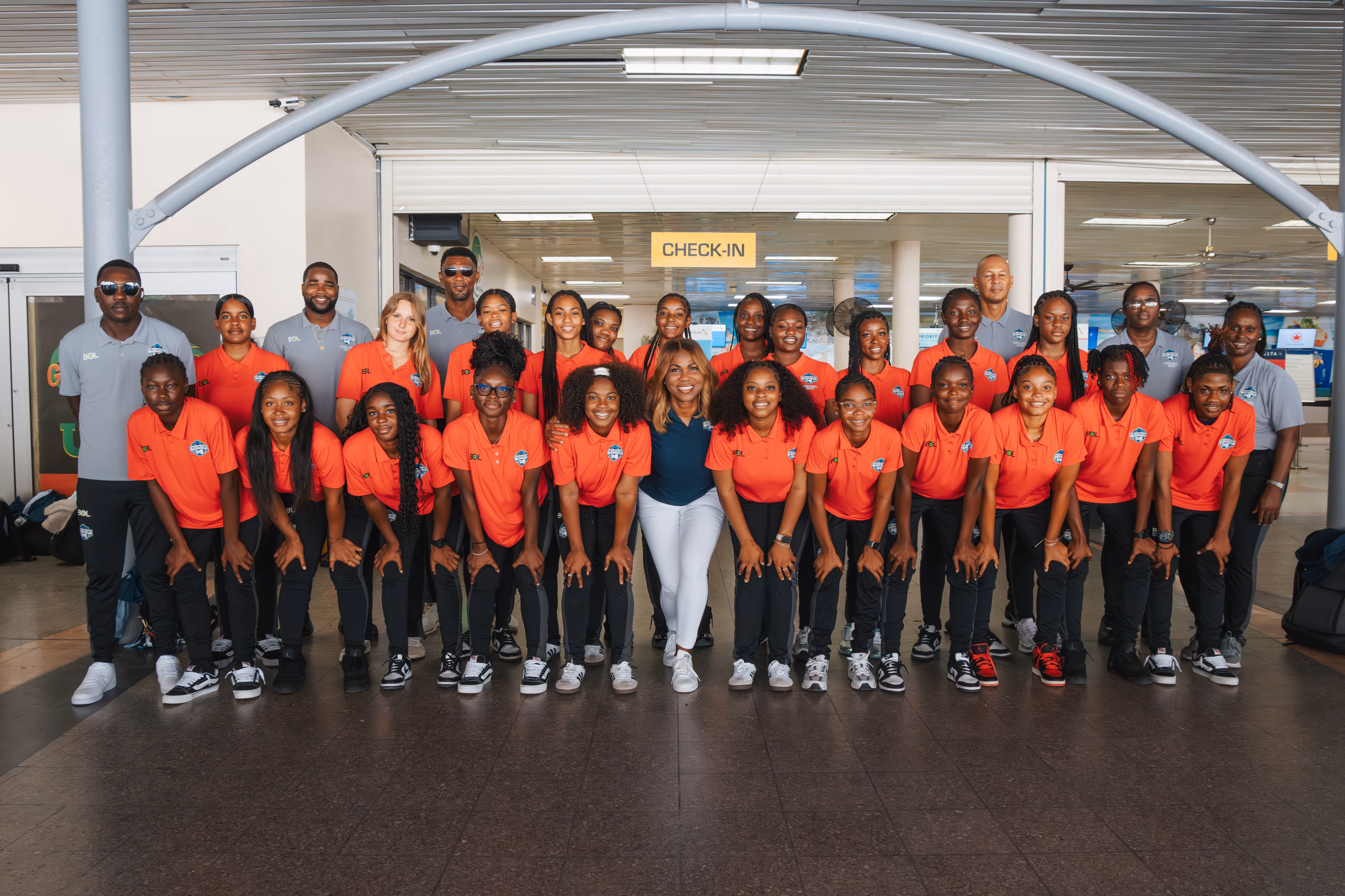 Group of young female athletes in orange jerseys and coaches in gray shirts posing at an indoor check-in area.