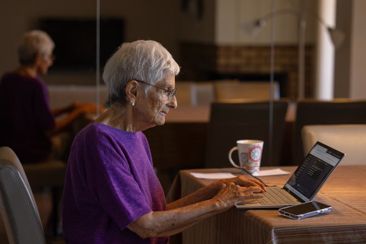 An older woman with short grey hair and glasses typing on a laptop at a table, with a cup of tea and a phone beside her and another person working in the background.