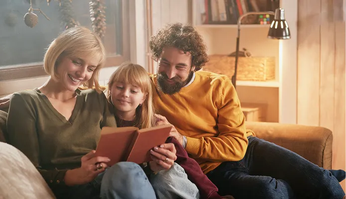 Eine glückliche Familie mit einer Frau, einem Mädchen und einem Mann sitzt auf einem Sofa und liest gemeinsam ein Buch in einem gemütlich beleuchteten Wohnzimmer.