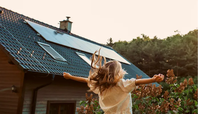 Ein Mädchen mit ausgestreckten Armen und wehenden Haaren steht im Garten vor einem Haus mit Solarpaneelen auf dem Dach bei warmem Sonnenlicht.