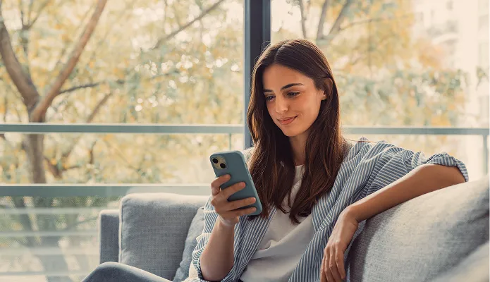 Eine Frau mit braunen Haaren sitzt entspannt auf einem grauen Sofa und schaut lächelnd auf ihr Smartphone, im Hintergrund sind Bäume mit Herbstlaub zu sehen.