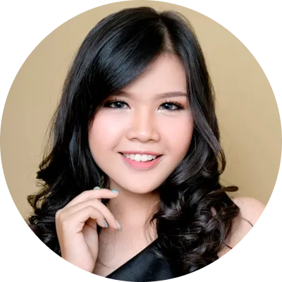 Portrait of a young woman with long, wavy black hair smiling gently, wearing black attire, and posing with her hand near her face against a neutral beige background.