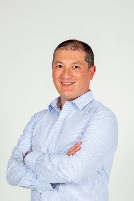 Smiling man with short dark hair wearing a light blue striped dress shirt with arms crossed standing against a plain light background.