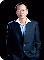 Man wearing glasses and a dark suit with a white shirt and blue tie posing with one hand in his pocket against a black background.